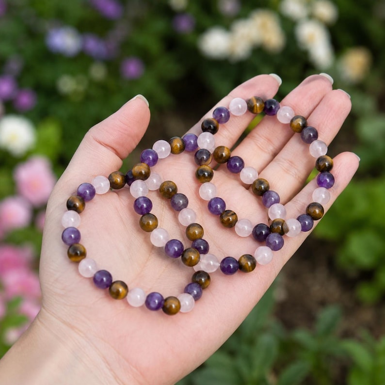 May include: Three stretch bracelets made of round beads in alternating colors of translucent light pink, deep purple, and golden brown tiger eye. The beads are strung on an elastic cord, creating a circular shape for each individual bracelet.