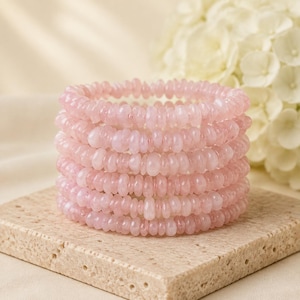 Puede incluir: Una pila de seis pulseras de cuarzo rosa claro con pequeñas cuentas lisas en forma de disco. Las piedras translúcidas de color rosa pálido están ensartadas en un patrón circular, creando una apariencia uniforme y delicada en cada pulsera.