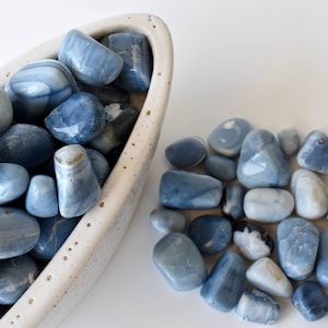 May include: A collection of blue and white tumbled stones in a white ceramic bowl and scattered on a white surface. The stones have a variety of shapes and sizes.