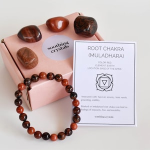 May include: A pink box containing a red, brown and orange crystal bracelet and four red, brown and orange crystals. The box has a sticker that says "soothing crystals". A white card with the text "ROOT CHAKRA (MULADHARA)" is next to the box. The card features a red symbol and text about the root chakra.