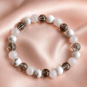 Puede incluir: Una pulsera circular de cuentas con cuentas redondas alternas en blanco s&oacute;lido, blanco y gris jaspeado, y marr&oacute;n oscuro transl&uacute;cido con inclusiones internas visibles. Las cuentas est&aacute;n ensartadas en un patr&oacute;n repetitivo formando un bucle.