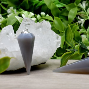 May include: Two gray agate crystal pendulums, one with a silver chain. The pendulums are pointed and have a smooth, polished finish. The larger pendulum is resting on a white crystal cluster.