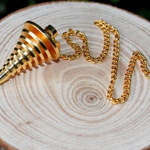 Op de afbeelding: Een gouden metalen slinger met een spiraalvormig ontwerp en een ketting. De slinger hangt aan een houten oppervlak.