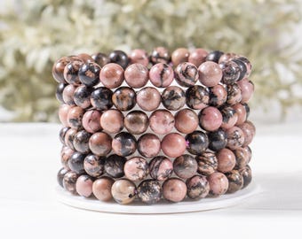 May include: A stack of five beaded bracelets made from round, polished rhodonite stones. The beads feature a mix of dusty pink and dark black mottled patterns, arranged in a cylindrical formation on a white circular base.