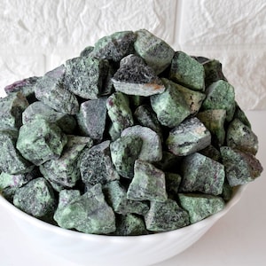 Könnte beinhalten: Eine weiße Schale gefüllt mit einem großen Haufen grüner und schwarzer, roher, unpolierter Edelsteine. Die Steine sind unregelmäßig geformt und haben ein geflecktes Aussehen.