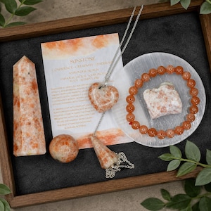 May include: A sunstone collection featuring a tall crystal tower, a sphere, a heart-shaped pendant on a silver chain, a pendulum, a beaded bracelet, and a raw stone piece on a white disc, accompanied by an informational card about sunstone.