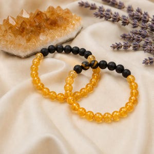 Könnte beinhalten: Zwei elastische Armbänder aus einer Kombination von runden, durchscheinenden gelben Perlen und porösen, mattschwarzen Lavaperlen. Die Armbänder sind in einem überlappenden kreisförmigen Muster angeordnet und zeigen den Kontrast zwischen den glatten gelben Steinen und den strukturierten schwarzen Vulkangesteinsperlen.