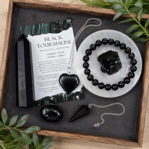May include: A black tourmaline crystal set including a polished tower, a heart-shaped pendant on a silver chain, a beaded bracelet, a raw stone, and a pointed pendulum on a chain, accompanied by an informational card about black tourmaline.