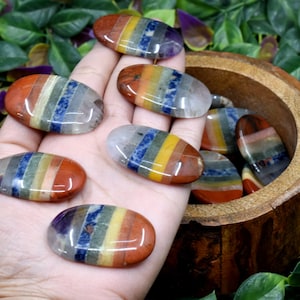 May include: A collection of seven chakra stones, each with a different colour, arranged in a rainbow pattern. The stones are smooth and polished, and they are displayed on a hand and in a wooden bowl.