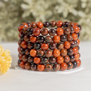 May include: A multi-strand bracelet composed of polished spherical beads in alternating shades of deep burnt orange, rust red, and dark chocolate brown. The beads feature natural mottled patterns and are arranged in a stacked, cylindrical formation.