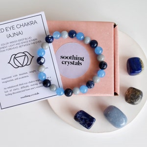 May include: A third eye chakra bracelet made with blue and grey stones, packaged in a pink box with a card that reads "Third Eye Chakra (Ajna)" and describes the chakra's properties. The card also includes the text "soothing crystals".