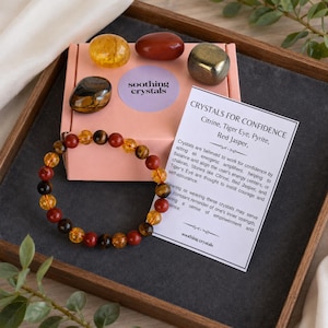 May include: A beaded bracelet featuring alternating yellow, brown, and red polished stones, presented with a pink box labelled soothing crystals, four loose tumbled stones, and an informational card titled Crystals for Confidence.