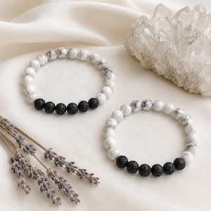 May include: Two stretch bracelets featuring round white marbled beads and a segment of five porous black lava beads. The bracelets are arranged on a soft fabric surface alongside a cluster of clear quartz crystals and dried lavender sprigs.