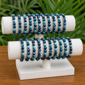 May include: A collection of beaded bracelets displayed on a white two-tier stand. Each bracelet consists of alternating round beads in shades of deep navy blue and vibrant turquoise, strung together in a repeating pattern on an elastic cord.
