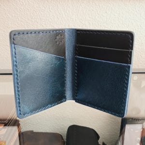 May include: A bi-fold wallet featuring a deep blue exterior and black interior pockets. The wallet is constructed with visible dark stitching along the edges and includes a small, embossed logo on the upper left interior panel.