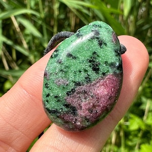 Könnte beinhalten: Ein grüner und rosafarbener Edelstein-Anhänger mit schwarzen Flecken. Der Anhänger ist oval geformt und hat eine glatte, polierte Oberfläche. Er ist an einer schwarzen Schnur befestigt.