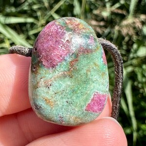 以下が含まれることがあります： 茶色の革紐に、緑とピンクの宝石のペンダント。宝石は、ピンクと緑の渦巻き模様で、まだら模様になっている。