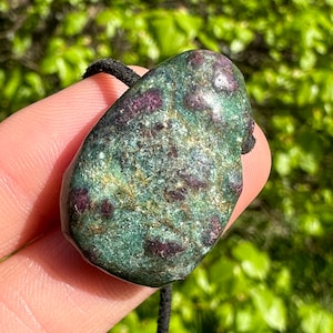Puede incluir: Un colgante de piedra preciosa verde y roja con un cordón negro. La piedra preciosa tiene una textura rugosa y natural y está salpicada de motas rojas y doradas.
