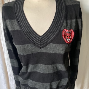 May include: A black and gray striped V-neck sweater with a devil heart patch. The sweater has long sleeves and a ribbed hem and cuffs. The devil heart patch is red with a smiling devil face.