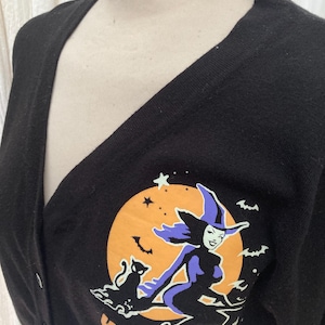 May include: Black cardigan with a V-neck and button closure. The front features a graphic of a witch on a broomstick in front of an orange moon, with a black cat and the text "KILLES STAR".