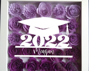 May include: A white graduation cap with the year 2022 and the name Meagan written below. The cap is displayed in a white frame with purple paper flowers.