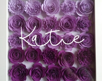 May include: A white frame with a purple and white ombre paper flower arrangement. The flowers are arranged in rows and the name "Katie" is written in white cursive lettering in the center of the frame.