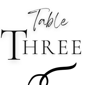 Op de afbeelding: Zwart-wit tafelnummerbord met de tekst "Table Three" en een decoratieve versiering.