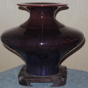 May include: A large, dark purple vase with a flared top and a bulbous body. The vase has a glossy finish and sits on a dark wooden stand. The color transitions from deep purple to a dark blue at the base.