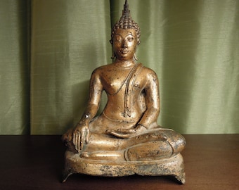 Op de afbeelding: Een gouden Boeddhabeeld dat in meditatieve houding zit met een serene uitdrukking. Het beeld staat op een voetstuk met een decoratief ontwerp.