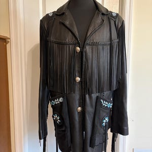 May include: A black leather jacket featuring long fringe detailing across the chest and sleeves. The jacket includes silver-toned metal buttons down the front and decorative patches with blue, white, and black beadwork on the shoulders and pockets.