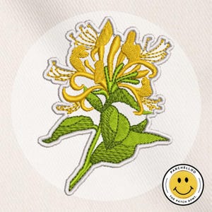Puede incluir: Parche bordado con una flor de madreselva amarilla con hojas y tallo verdes, contorneada en blanco. El parche está sobre un fondo blanco con forma circular. En la esquina inferior derecha hay un logotipo circular con una cara sonriente y el texto "PARCHELLOO THE PATCH DPMT".