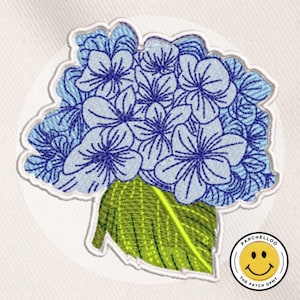 Puede incluir: Parche bordado con un grupo de flores de hortensia azules con una hoja verde. Las flores tienen costuras detalladas y están sobre un fondo blanco. El parche tiene un borde blanco y un pequeño logotipo en la esquina inferior derecha.