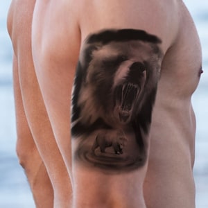 Peut inclure: Un homme avec un tatouage d'un ours sur le haut de son bras. L'ours rugit la gueule ouverte et a un design sombre et détaillé.