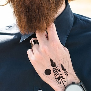 May include: A man with a long beard and a black shirt is wearing a temporary tattoo on his hand. The tattoo is a black and white design of a forest with trees and a circle.