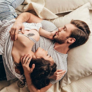 May include: A couple lying in bed together, the woman is wearing a striped shirt and the man is wearing a grey t-shirt. The woman has a feather tattoo on her chest.