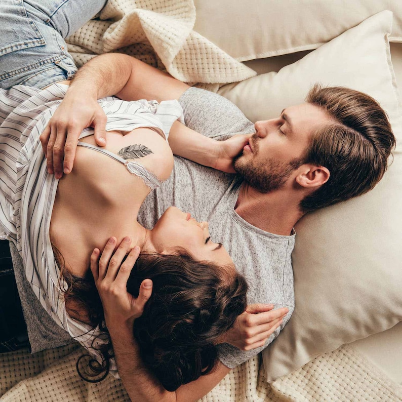 May include: A couple lying in bed together, the woman is wearing a striped shirt and the man is wearing a grey t-shirt. The woman has a feather tattoo on her chest.