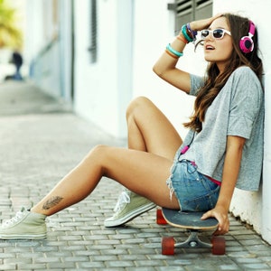 May include: A young woman wearing a grey shirt, denim shorts, and pink headphones sits on a skateboard with her legs extended. She is wearing sunglasses and has a feather tattoo on her ankle.