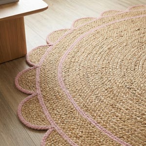 May include: A round jute rug with a scalloped edge and a pink border. The rug is made of woven natural fibers and has a neutral color palette. The rug is placed on a wooden floor.