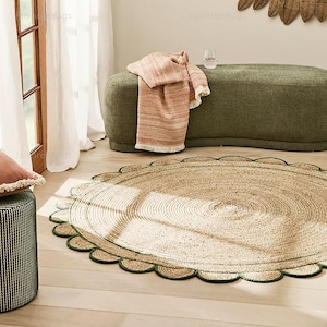 May include: A round jute rug with a scalloped edge, featuring a natural tan color with a green border. The rug is placed on a light wood floor, with a green upholstered bench and a pink throw blanket in the background.