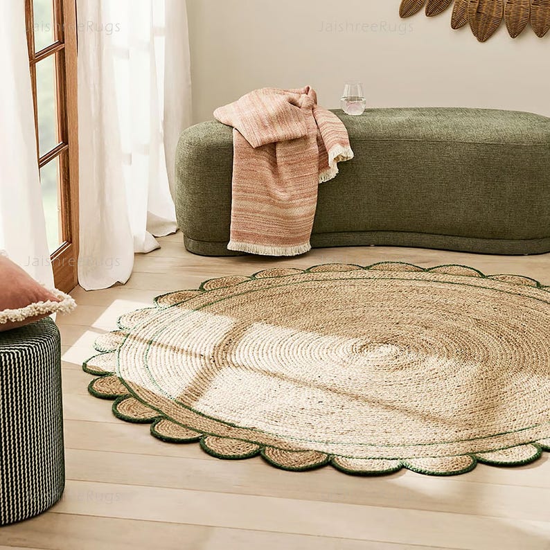 May include: A round jute rug with a scalloped edge, featuring a natural tan color with a green border. The rug is placed on a light wood floor, with a green upholstered bench and a pink throw blanket in the background.