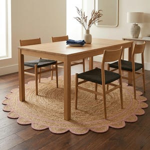 May include: A light wood dining table with four matching chairs. The chairs have black woven seats and light wood frames. A round jute rug with a pink scalloped edge sits beneath the table and chairs. A vase of dried flowers sits on the table.