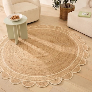May include: A round, natural jute rug with a scalloped edge. The rug features a light brown color with a white border and a textured, woven design. The rug is placed on a light-colored wooden floor, with furniture in the background.