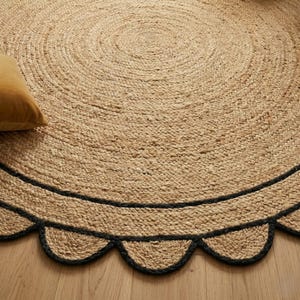 May include: A round jute rug with a scalloped black border. The rug has a natural, woven texture and a light brown color. A yellow pillow is visible in the upper left corner. The rug is placed on a wooden floor.