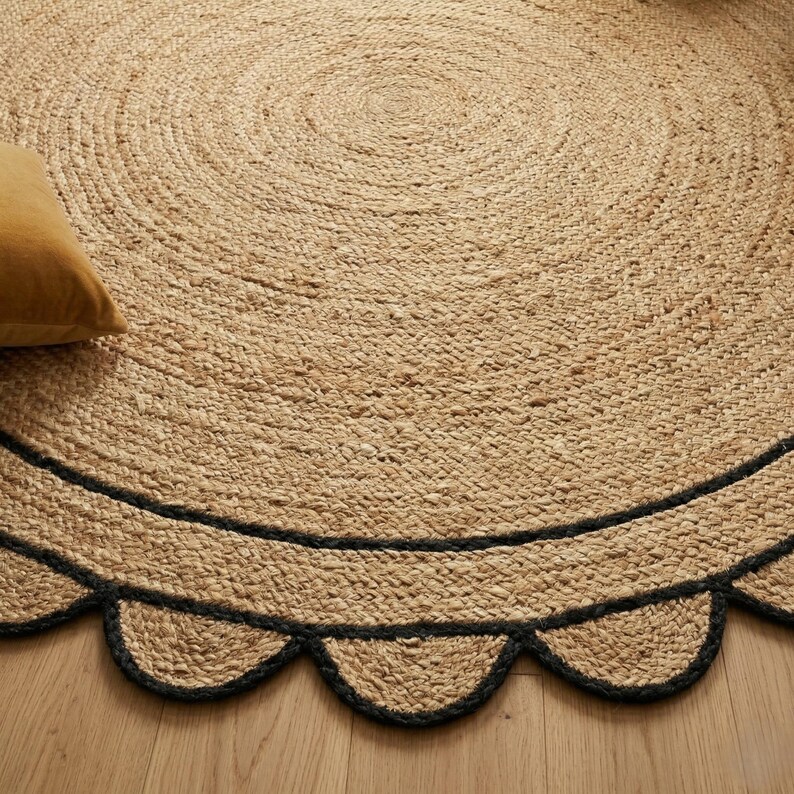 May include: A round jute rug with a scalloped black border. The rug has a natural, woven texture and a light brown color. A yellow pillow is visible in the upper left corner. The rug is placed on a wooden floor.