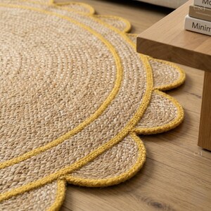 May include: A round jute rug with a scalloped edge, featuring a yellow border. The rug is made of woven natural fibers and sits on a wooden floor. The rug's design adds a touch of texture and warmth to any space.