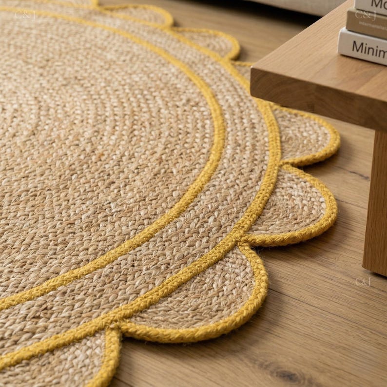 May include: A round jute rug with a scalloped edge, featuring a yellow border. The rug is made of woven natural fibers and sits on a wooden floor. The rug's design adds a touch of texture and warmth to any space.
