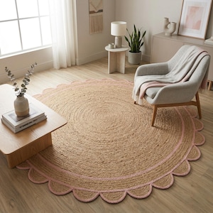 May include: A round jute rug with a scalloped pink border. The rug is made of natural fibers and has a woven texture. The rug is placed on a wooden floor in a living room setting.