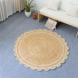 May include: Round jute rug with a scalloped edge and a white border. The rug has a natural, light brown color and is placed on a blue-gray floor. The rug's design adds a touch of texture and warmth to the room.