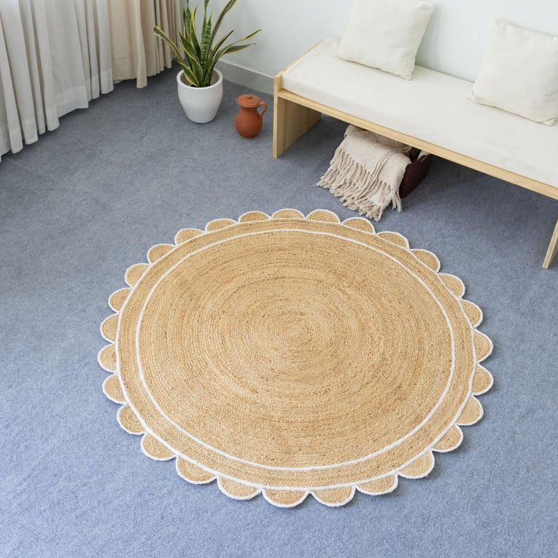 May include: Round jute rug with a scalloped edge and a white border. The rug has a natural, light brown color and is placed on a blue-gray floor. The rug's design adds a touch of texture and warmth to the room.