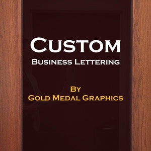 Op de afbeelding: Een bord met de tekst "CUSTOM BUSINESS LETTERING BY GOLD MEDAL GRAPHICS" in witte letters op een donkerbruine achtergrond.